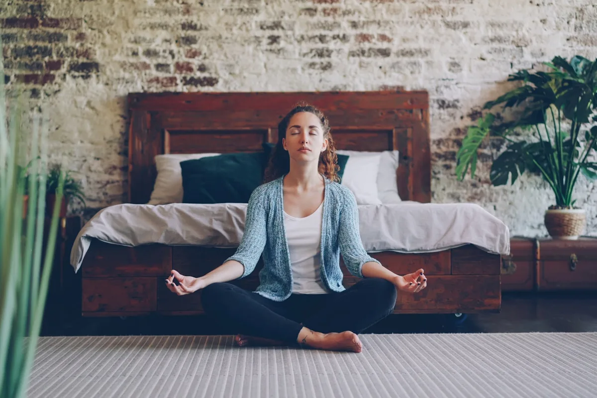 Nő lótuszülésben, csukott szemmel meditál egy meleg, növényekkel teli szobában
